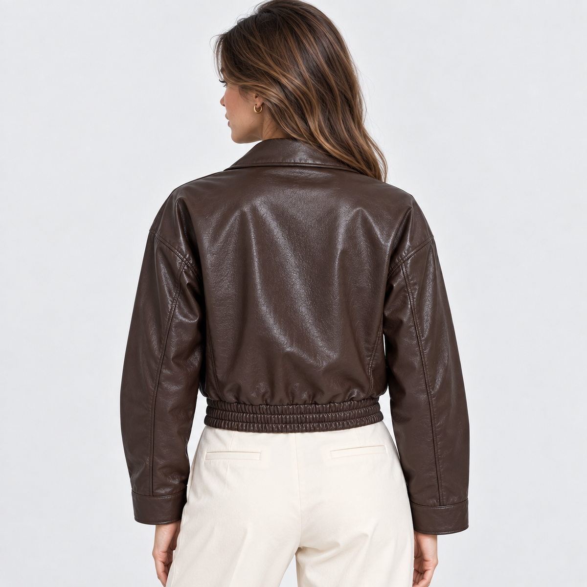 A woman shown from the back wearing a dark brown faux leather cropped jacket with a gathered elastic hem, paired with cream-colored pants, against a plain light gray background.