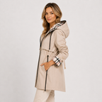 A woman modeling a beige hooded trench coat with black drawstrings and a plaid lining visible on the hood and cuffs, paired with light beige pants against a plain light gray studio background.