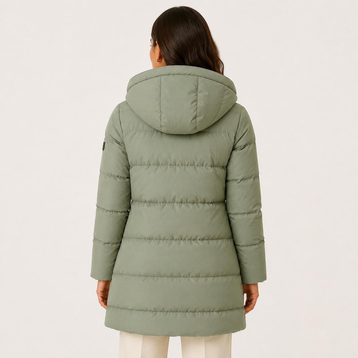 A woman viewed from the back wearing a sage green hooded puffer coat, paired with off-white pants, against a plain light background.