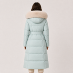 A model viewed from the back wearing a long, light blue-green puffer coat with a beige faux-fur trimmed hood and a matching waist belt, set against a solid light beige background.