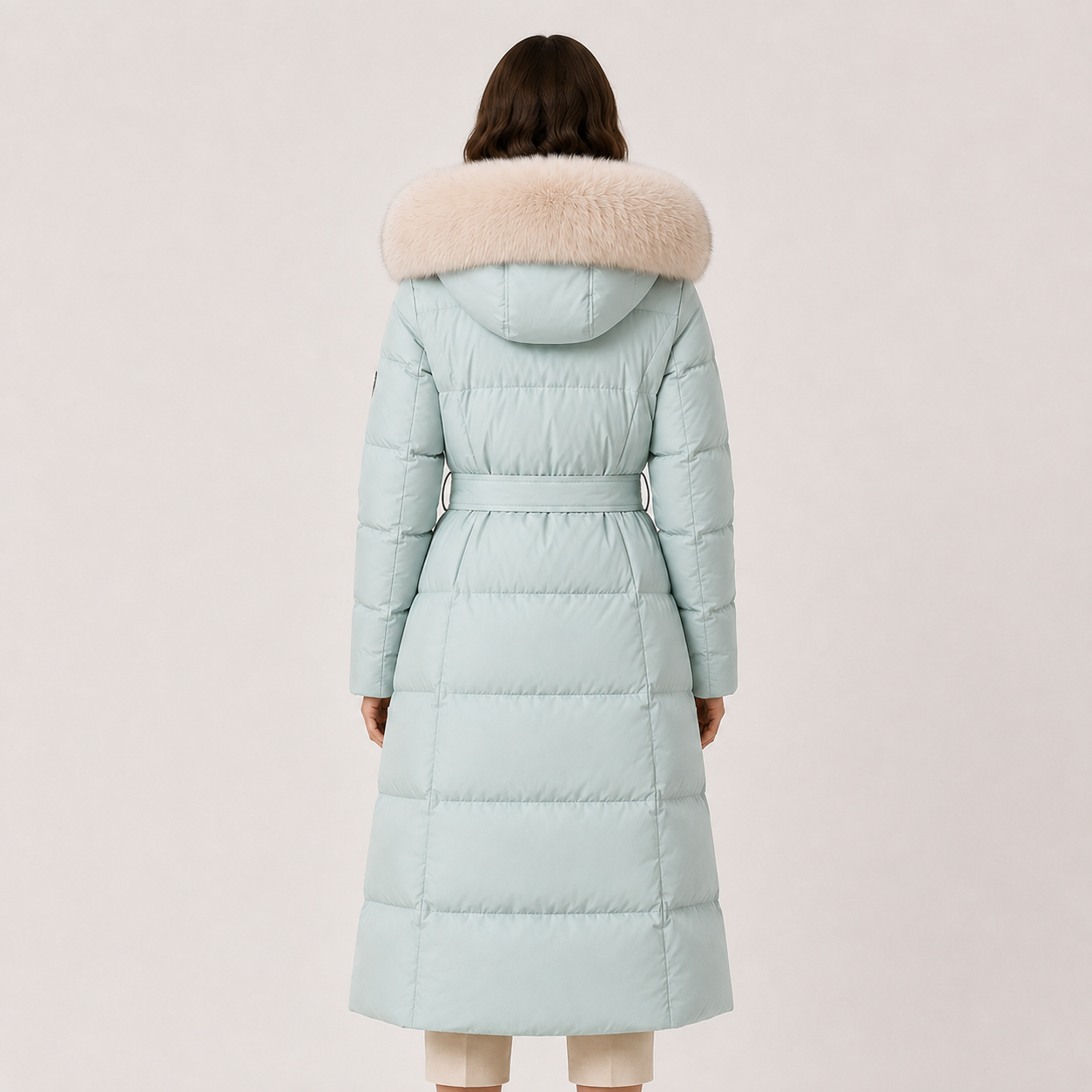 A model viewed from the back wearing a long, light blue-green puffer coat with a beige faux-fur trimmed hood and a matching waist belt, set against a solid light beige background.