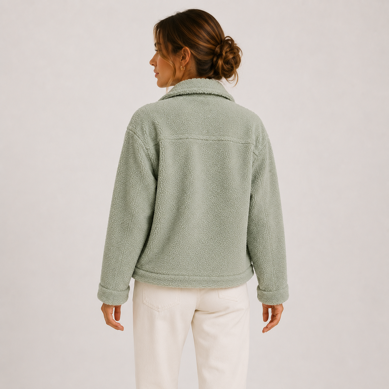 A woman shown from the back wearing a light sage green sherpa fleece jacket and white pants, standing against a plain light gray studio background.
