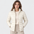 A woman modeling a short, quilted puffer jacket in a warm off-white/cream color, worn over a matching cream sweater and beige trousers, against a light gray studio background.