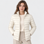 A woman modeling a short, quilted puffer jacket in a warm off-white/cream color, worn over a matching cream sweater and beige trousers, against a light gray studio background.