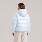 A woman shown from the back wearing a shiny light blue puffer jacket and cream pants, photographed against a plain light gray studio background.