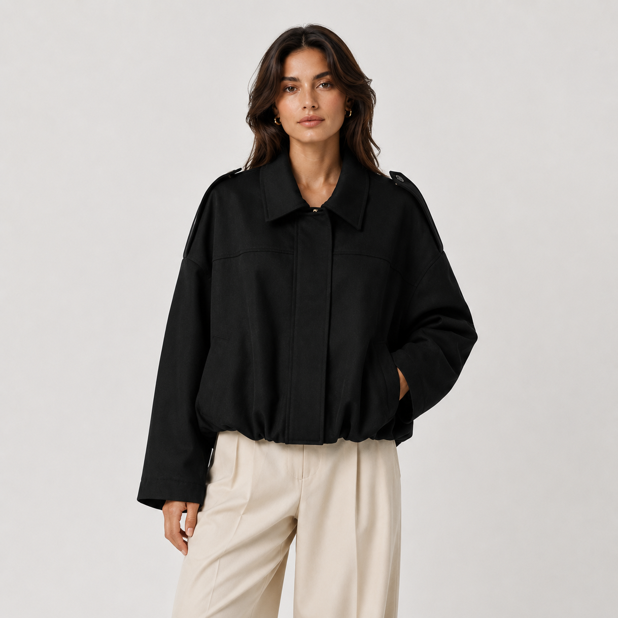 A woman modeling a black cropped jacket with shoulder epaulets and front pockets, paired with light beige trousers, against a plain light gray studio background.
