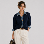 A woman models a navy blue knit button-up shirt with a light blue denim collar and cuffs, paired with cream trousers and a brown belt, against a light gray studio background.