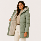 A woman models a sage green hooded puffer jacket with a beige sherpa lining, styled with a beige top and white pants against a plain white studio background.