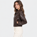 A woman wearing a dark brown faux-leather cropped jacket and cream-colored trousers, shown from a side profile against a plain white studio background.