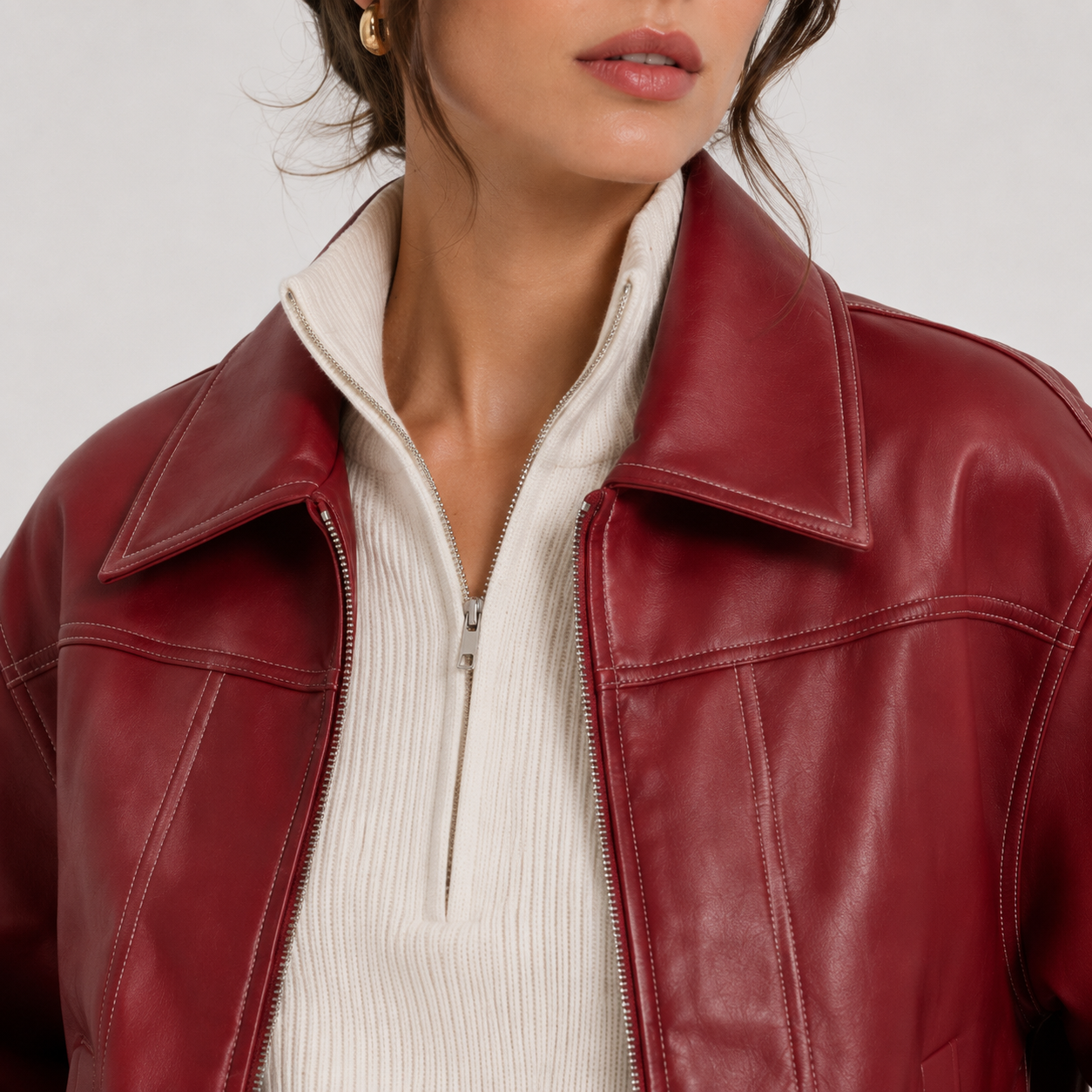 A close-up of a model wearing a deep burgundy leather jacket over a cream ribbed half-zip sweater, against a plain light gray background.
