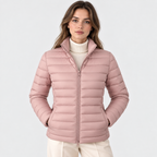A woman wearing a light pink puffer jacket over a cream ribbed turtleneck sweater and cream trousers, photographed against a clean light gray studio background.
