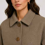 Close-up of a woman wearing a textured taupe-brown coat with a pointed collar and two brown buttons, paired with small gold hoop earrings against a light gray background.