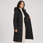 A woman modeling a long black hooded puffer coat over a cream turtleneck and cream pants, standing against a light gray studio background.