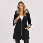 A woman modeling a black hooded coat with a plaid patterned hood lining and matching plaid cuffs, worn over a white top and black pants against a light gray studio background.