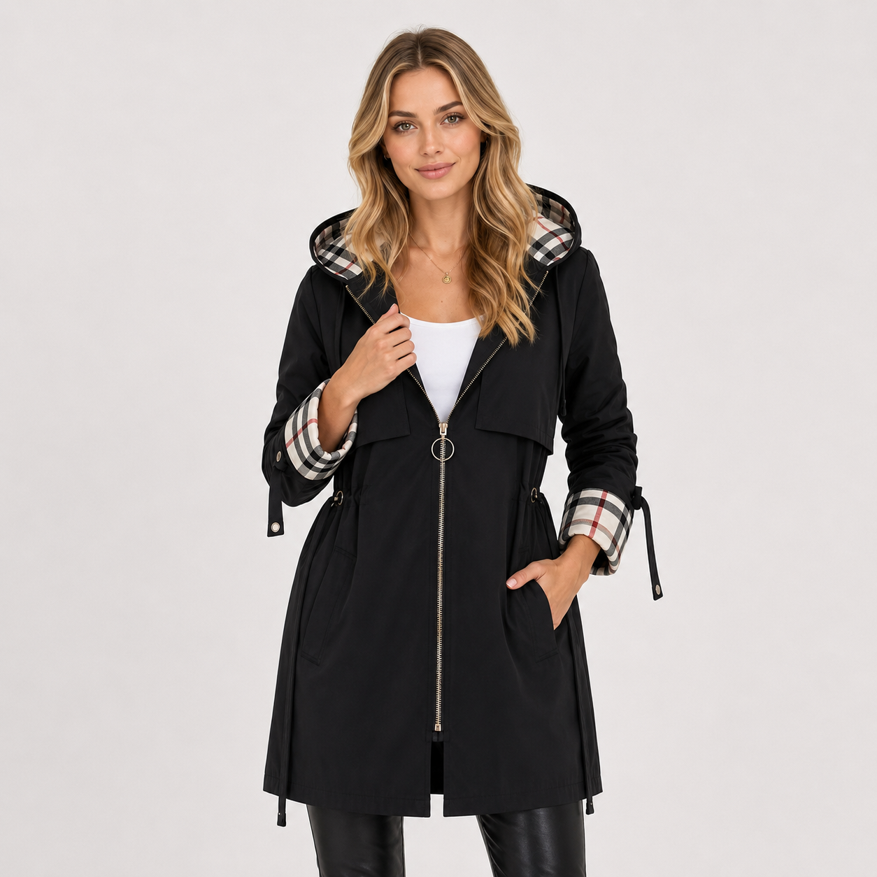 A woman modeling a black hooded coat with a plaid patterned hood lining and matching plaid cuffs, worn over a white top and black pants against a light gray studio background.