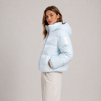 A woman wearing a shiny light blue puffer jacket with a high collar and white pants, standing against a plain light gray studio background.