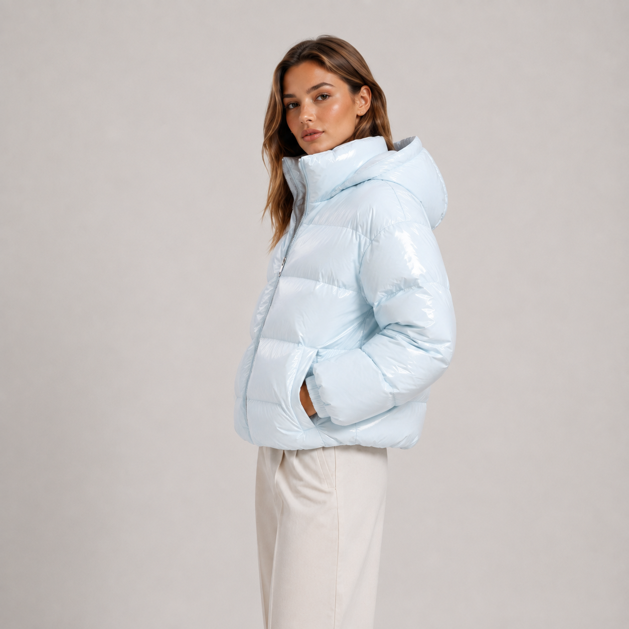 A woman wearing a shiny light blue puffer jacket with a high collar and white pants, standing against a plain light gray studio background.