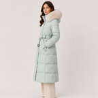 A woman modeling a long, light mint green puffer coat with a large beige faux-fur trimmed hood and a matching tie belt, set against a plain light beige studio background.