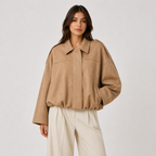 A woman modeling an oversized tan faux-suede jacket with a relaxed fit, wide lapels, and front flap pockets, paired with cream wide-leg trousers, against a light gray studio background.