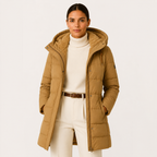 A woman modeling a mid-length tan hooded puffer jacket with a soft sherpa-lined hood, worn over a cream turtleneck and cream trousers with a brown belt, against a plain light background.