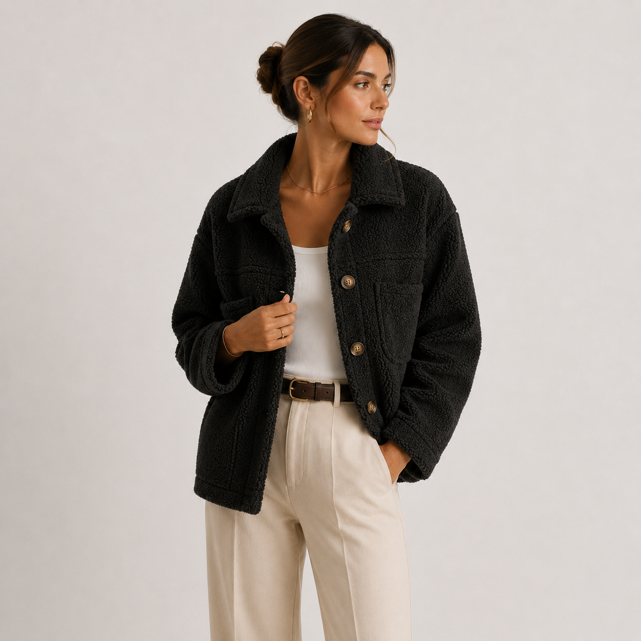 A woman wearing a dark charcoal textured jacket over a white top and cream trousers, standing against a light gray studio background.