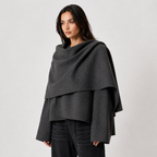 A woman modeling a dark gray draped wrap or poncho-style outer layer over a black top and black pants, photographed against a plain light gray studio background.