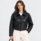 A woman modeling a black faux-leather cropped jacket with a zip front and collar, paired with cream trousers, against a light gray studio background.