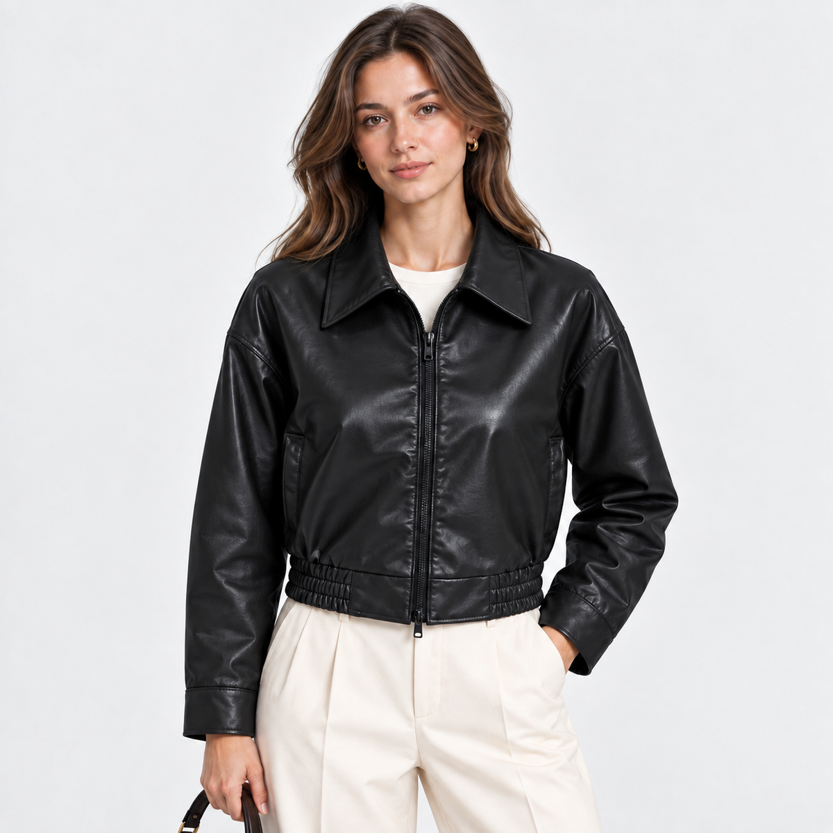 A woman modeling a black faux-leather cropped jacket with a zip front and collar, paired with cream trousers, against a light gray studio background.