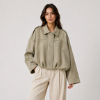 A woman modeling an oversized light olive green suede jacket with a collar and front zipper, paired with cream wide-leg trousers, against a plain light gray studio background.