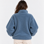 A woman viewed from behind wearing a dusty blue sherpa fleece jacket and light beige pants against a plain white background.