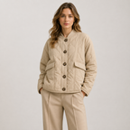 A woman modeling a light beige quilted jacket with dark buttons and matching beige trousers, photographed against a neutral light gray studio background.