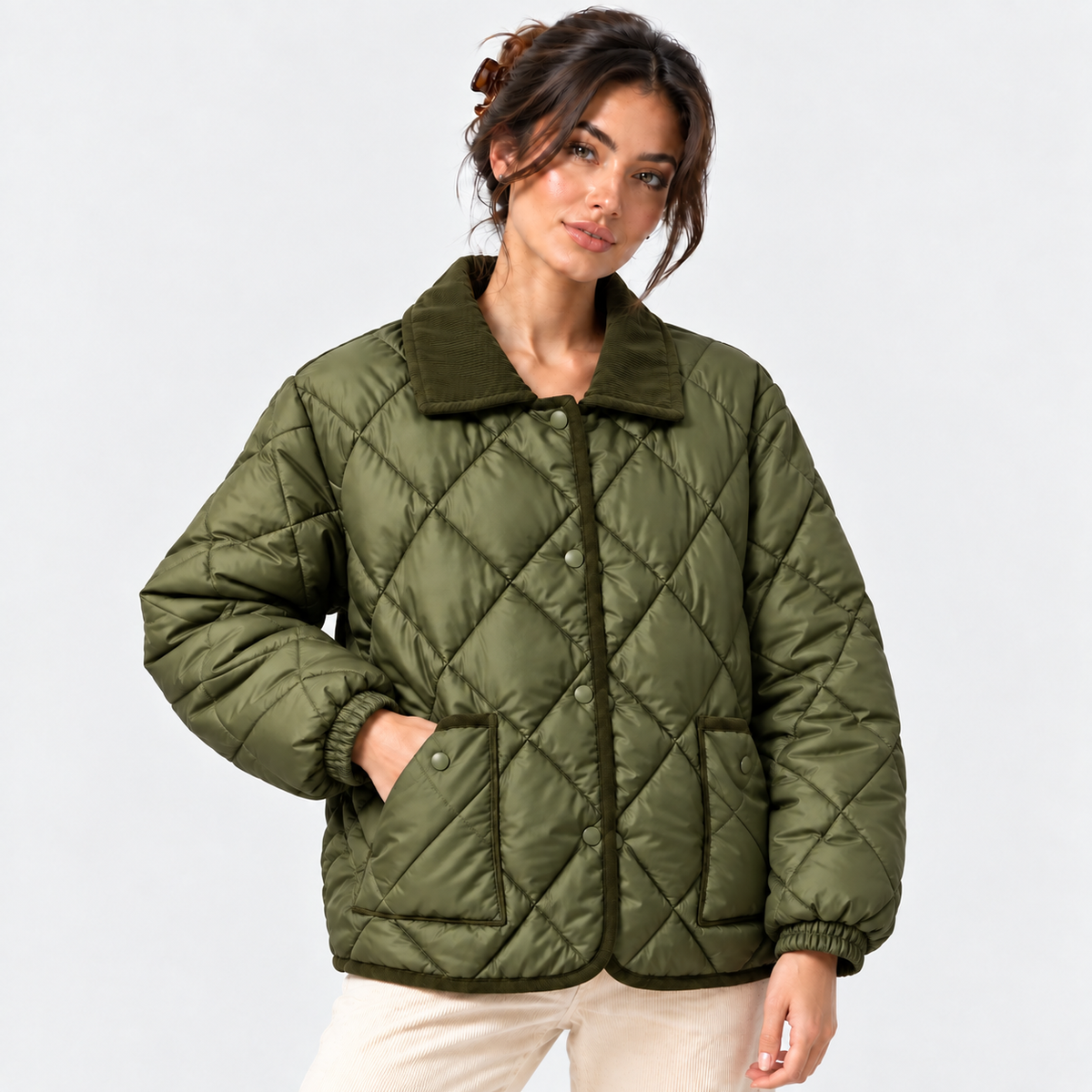 A woman wearing an olive green diamond-quilted puffer jacket with a corduroy-style collar and front buttons, paired with light beige pants, against a plain light gray studio background.