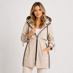 A woman modeling a beige hooded raincoat with a black front zipper, black drawstrings, and a plaid pattern lining the hood and cuffs, paired with white pants against a light gray studio background.