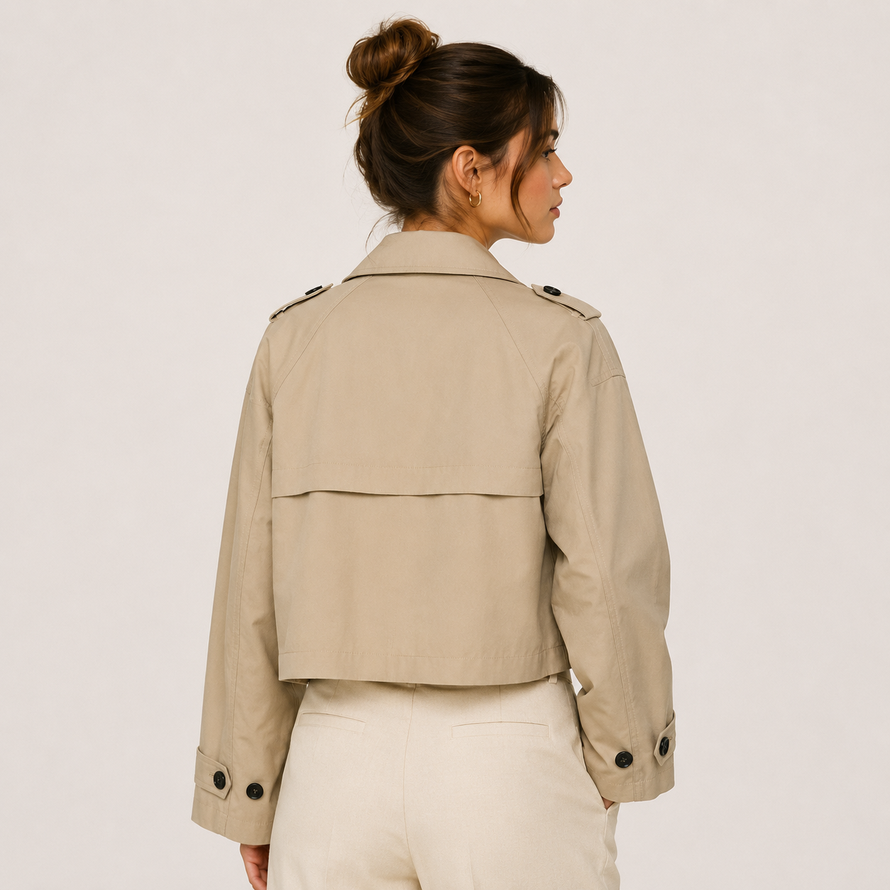A woman shown from the back wearing a cropped beige trench-style jacket with shoulder epaulets and button details, paired with light cream pants, against a plain light background.