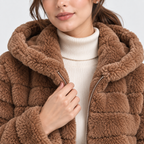 A close-up of a woman wearing a chunky brown sherpa fleece jacket with a large plush hood over a cream ribbed turtleneck sweater.