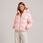 A woman wearing a shiny light pink puffer jacket with a hood and front zipper, paired with light beige pants, photographed against a plain light gray studio background.