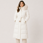 A woman wearing a long white puffer coat with a large fluffy fur-trimmed hood and a matching waist tie, photographed against a plain light studio background.