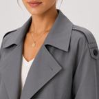 Close-up of a woman wearing a gray trench coat over a white top, with a delicate gold pendant necklace visible against a light gray background.