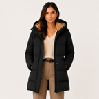 A woman wearing a black hooded puffer jacket with a tan sherpa-lined hood, styled over a cream ribbed sweater and tan trousers, photographed against a plain light gray studio background.