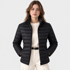 A woman modeling a black puffer jacket over a cream sweater and cream trousers, photographed against a light gray studio background.