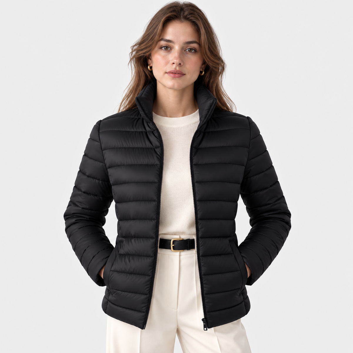 A woman modeling a black puffer jacket over a cream sweater and cream trousers, photographed against a light gray studio background.