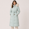 A woman wearing a long mint green puffer coat with a large beige faux-fur collar and a matching tie belt, standing against a light beige studio background.