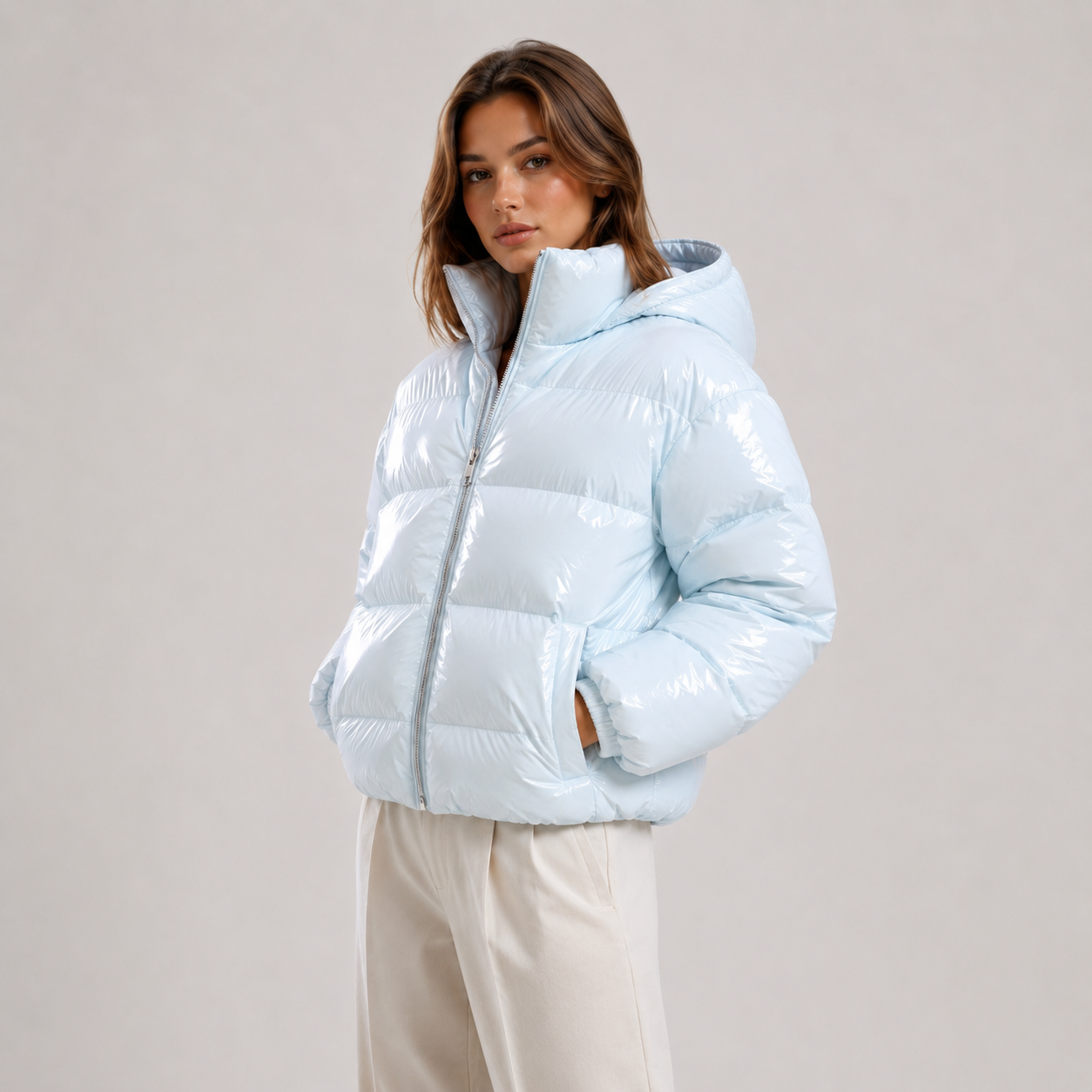 A woman wearing a shiny light blue puffer jacket with a high collar and front zipper, paired with cream-colored pants, photographed against a neutral light gray studio background.