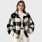 A woman modeling a black and off-white buffalo plaid fuzzy faux fur jacket over a white ribbed turtleneck sweater and cream trousers, shot against a solid light gray background.