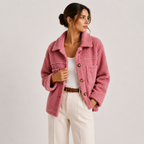 A woman wearing a dusty pink sherpa fleece jacket with brown buttons, styled with a white tank top and cream-colored trousers, photographed against a light gray studio background.