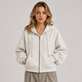 A woman wearing an oversized light beige zip-up hoodie with drawstrings and front pockets, paired with taupe pants, photographed against a plain light gray studio background.