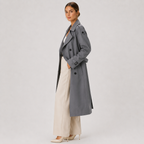A woman modeling a long double-breasted gray trench coat over a white top and cream wide-leg trousers, standing against a light gray studio background.