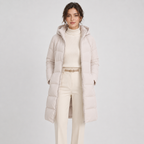 A woman modeling a long, light beige hooded puffer coat over a cream turtleneck sweater and matching cream trousers, photographed against a clean light gray studio background.