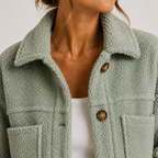 Close-up of a person wearing a sage green sherpa fleece button-up jacket over a white ribbed top, with a delicate gold chain necklace visible.