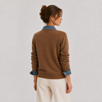 A woman shown from the back wearing a brown knit sweater with rolled-up sleeves revealing a blue denim collar and cuffs, paired with cream pants, against a light gray studio background.